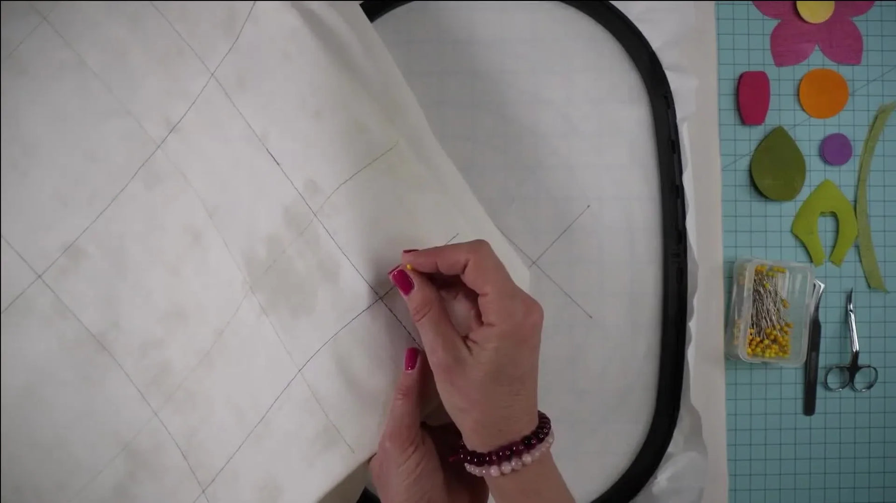 A close-up shows a pin being inserted straight down through the fabric’s marked intersection to create a pivot point for precise hoop alignment.