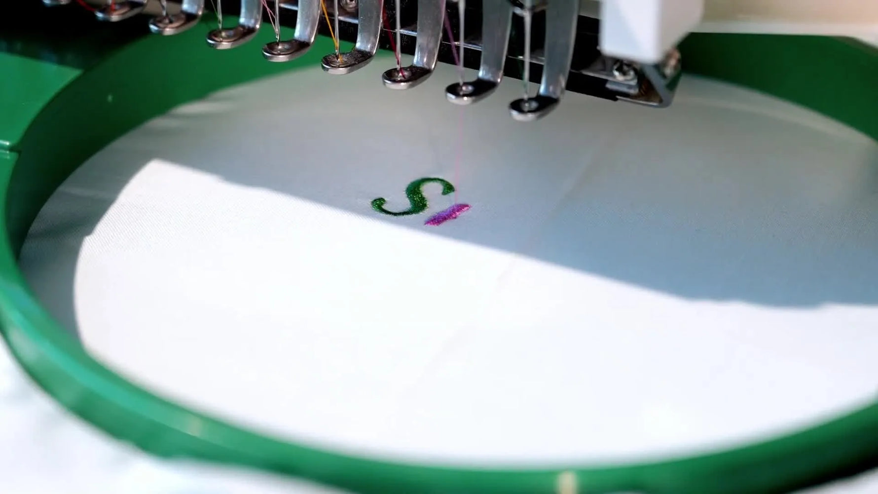 A close-up of a Smartstitch multi-needle machine stitching a green letter onto white fabric inside a round tubular hoop.
