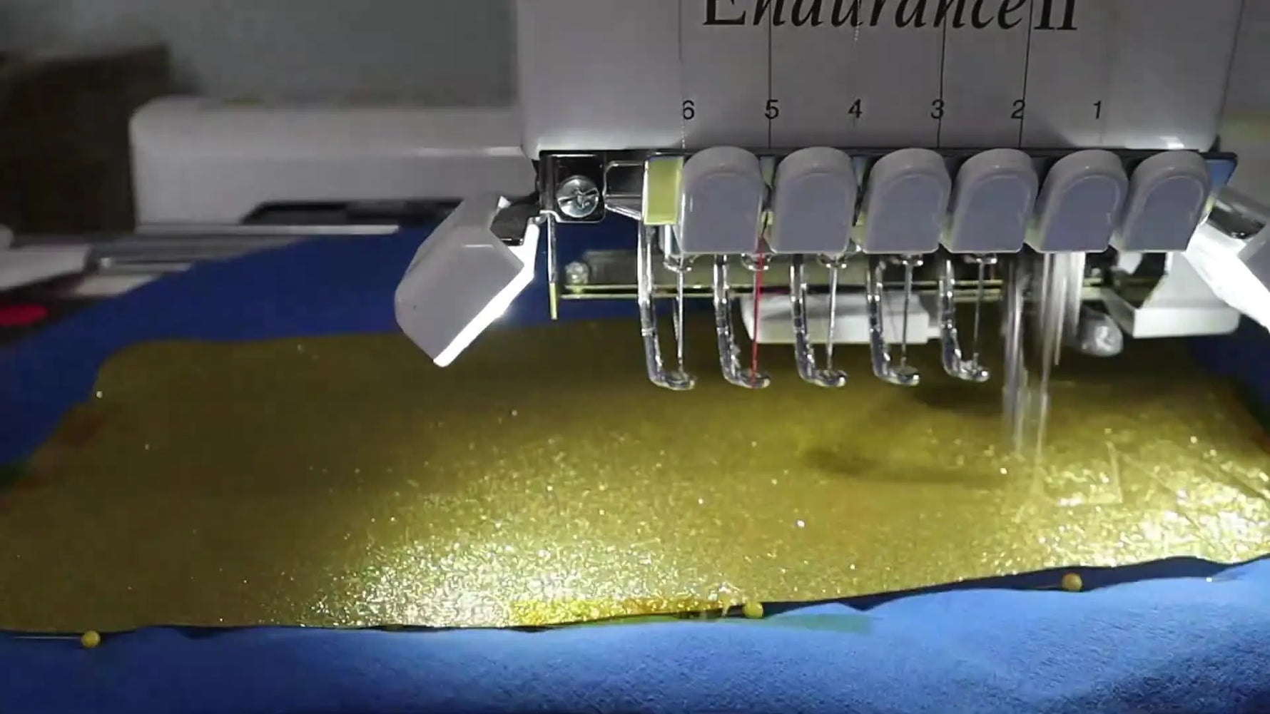 A close-up of a multi-needle embroidery machine stitching an outline through gold glitter HTV on a blue fleece blanket.