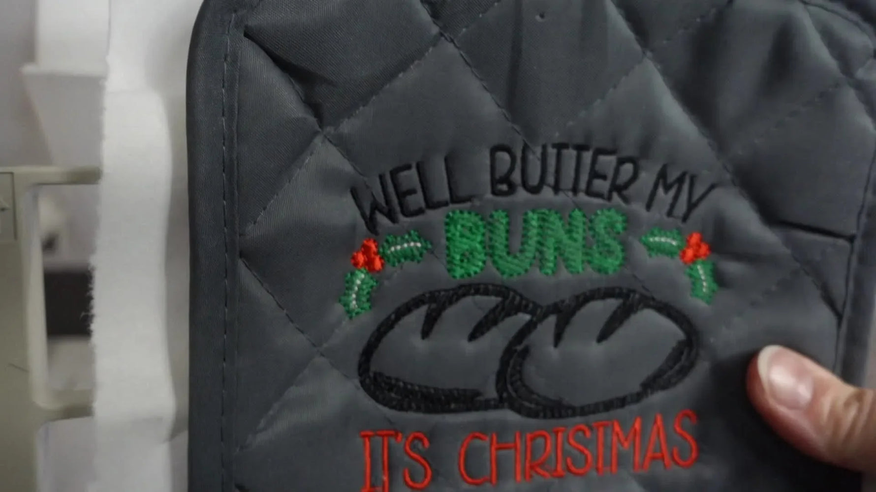 A close-up of the finished embroidered pot holder still mounted in the hoop on the embroidery machine, showing crisp lettering and clean stitch coverage.