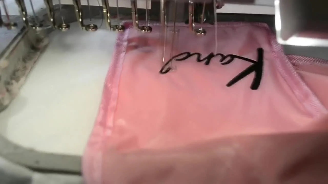 A close-up of the Brother PR1000e actively stitching black lettering onto a pink Velcro wallet that has been floated on stabilizer in a 5x7 hoop.