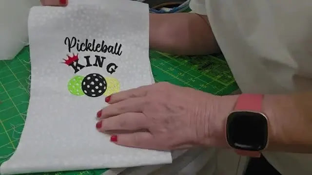 The finished “Pickleball King” embroidery is held up to show crisp lettering, colorful pickleballs, and a clean final result.