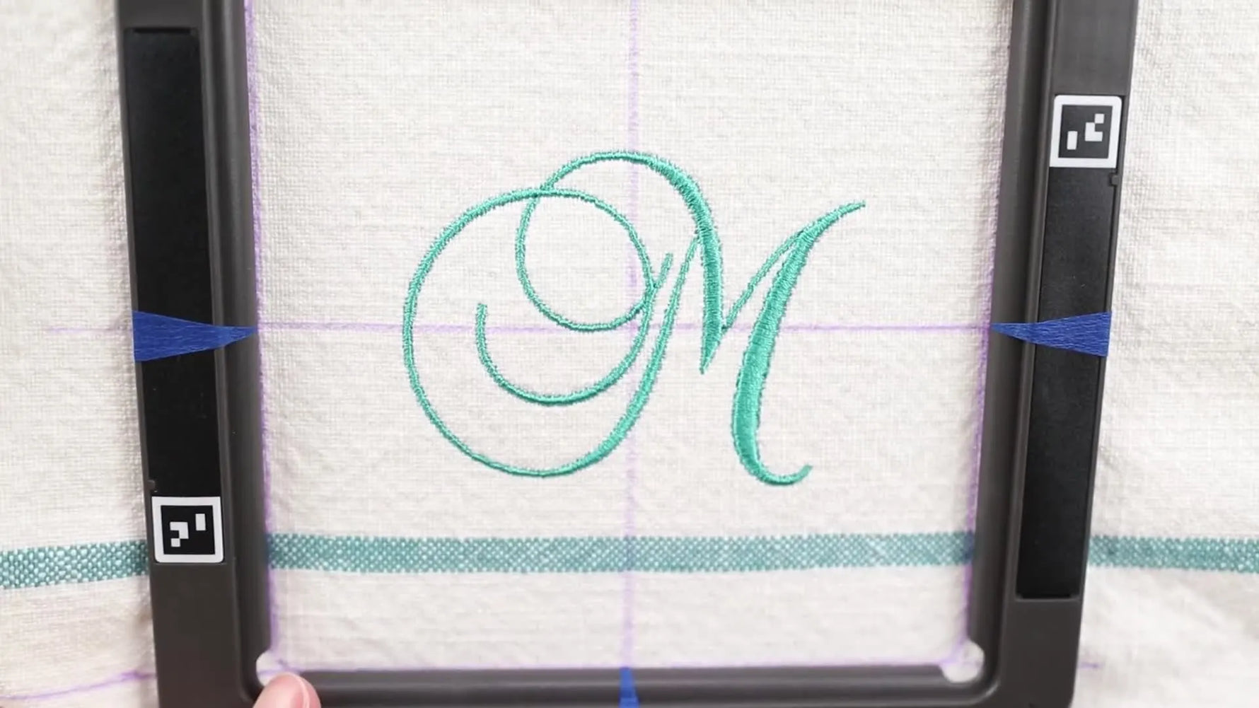 A finished teal “M” monogram stitched neatly on a white cotton napkin while still held in the magnetic hoop.