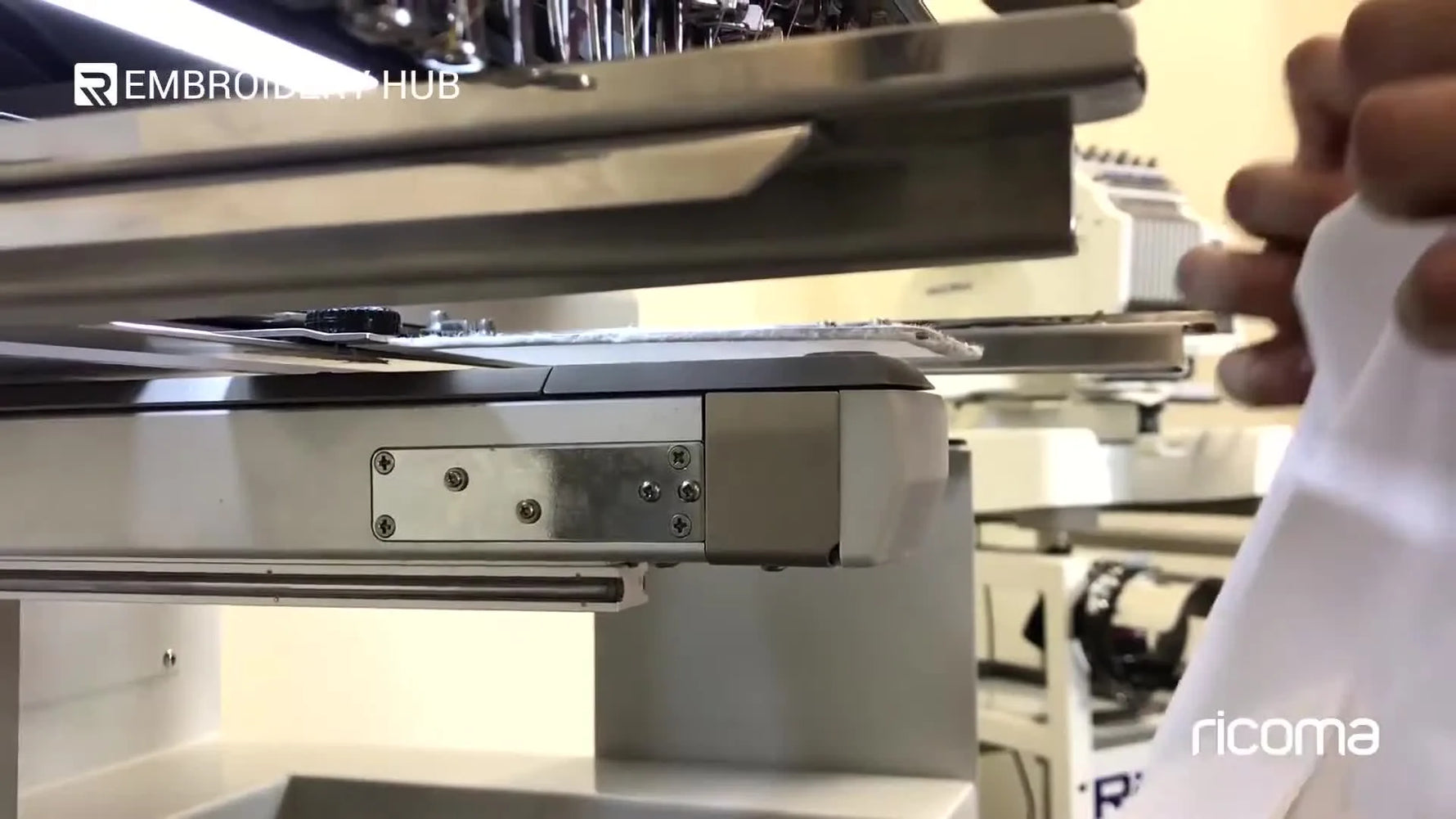 A close-up shows a dress shirt pocket being carefully floated onto a narrow rectangular frame arm mounted on a multi-needle embroidery machine.
