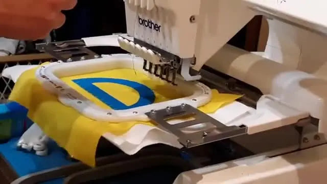 Overhead view of a Brother PR-series machine stitching a satin border on a blue appliqué letter while the MaggieFrame magnetic hoop holds the base fabric flat and stable.