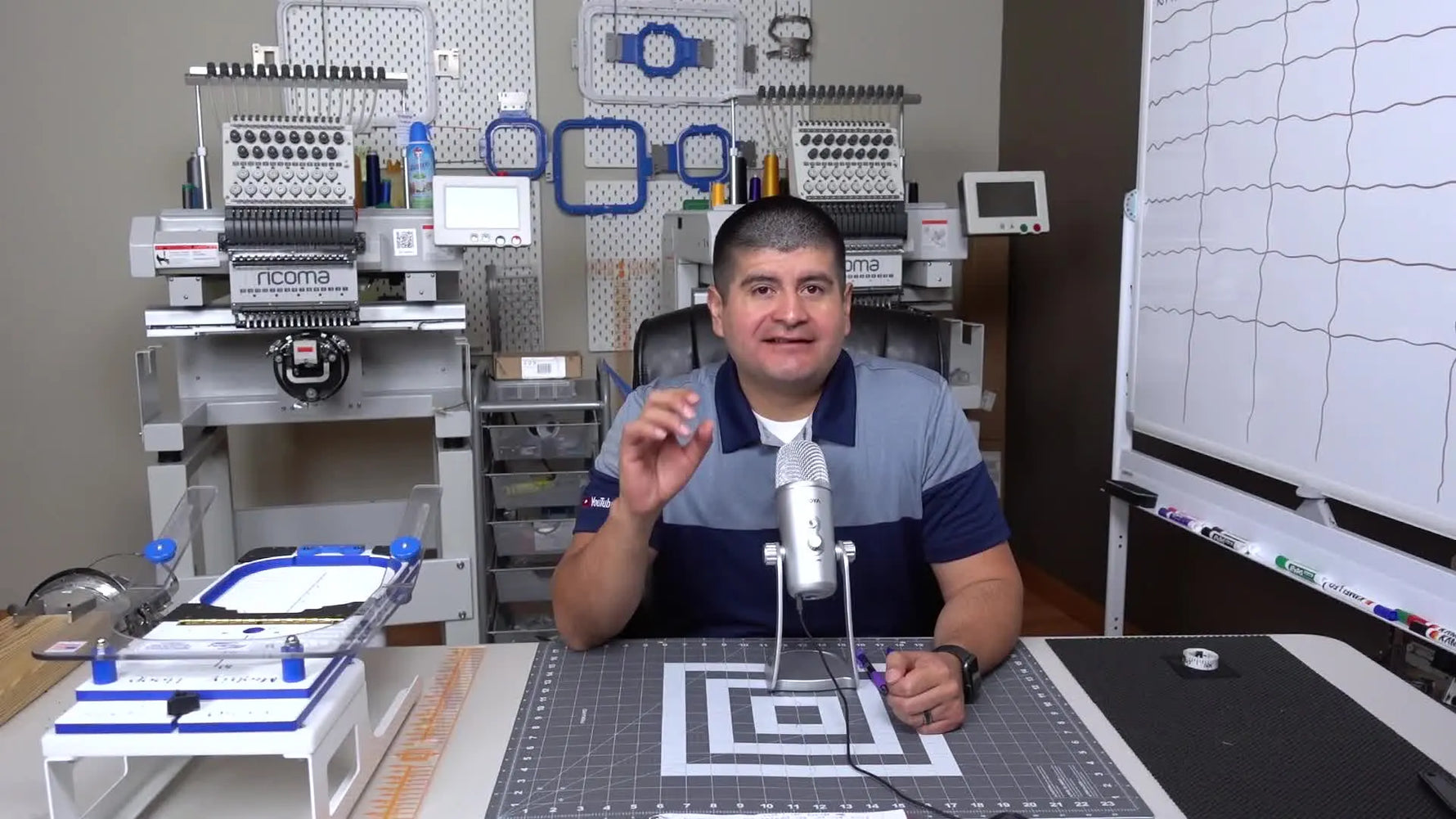 A digitizing instructor sits at a work desk with a multi-needle embroidery machine behind him and magnetic hoops laid out in front, setting the tone for a practical, production-focused lesson.