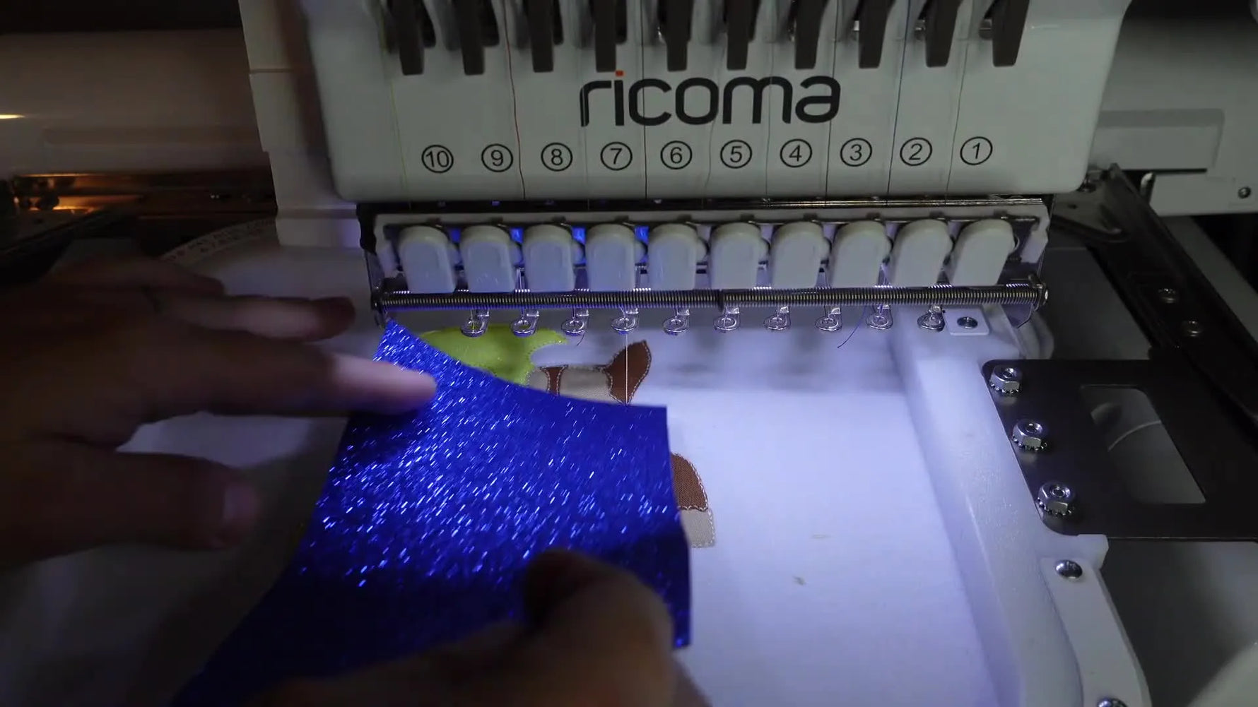 A multi-needle embroidery machine stitching the details of the Chase character appliqué onto the shirt, showing blue and brown fabric pieces.