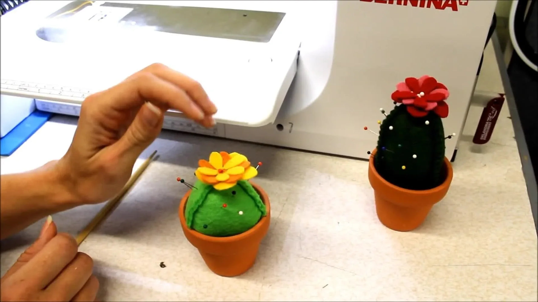 Three finished in-the-hoop cactus pincushions sit in small terracotta pots with pins inserted, showing the final 3D shape and flower topper.