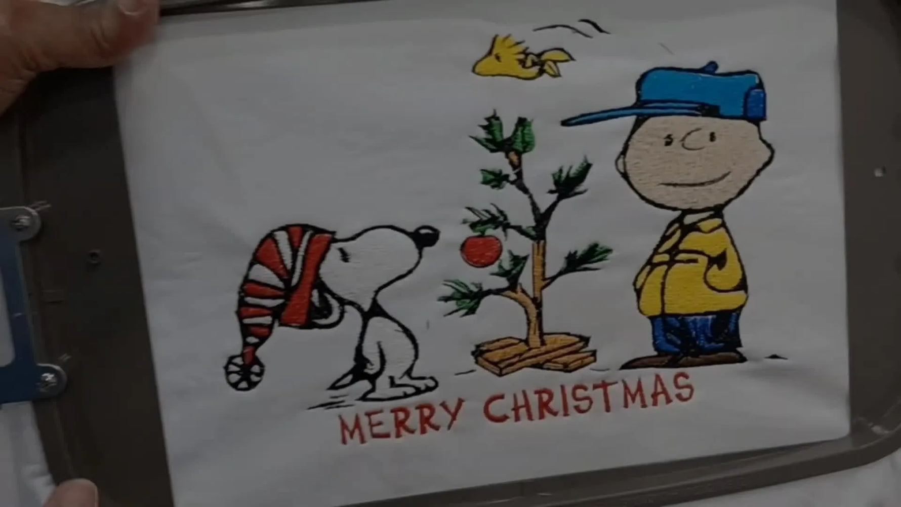 The finished Charlie Brown Christmas embroidery is held up in the hoop, showing clean stitching and balanced placement on the hoodie front.