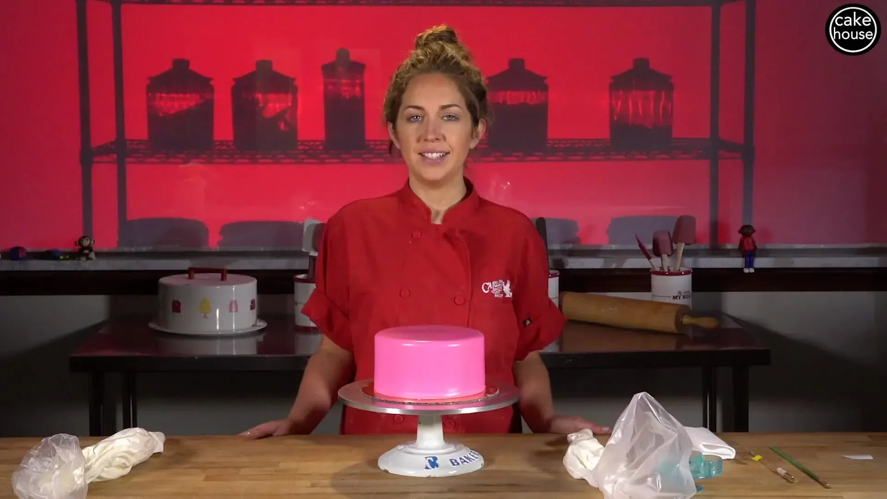 A woman in a red chef's coat standing behind a pink fondant-covered cake, introducing the tutorial.
