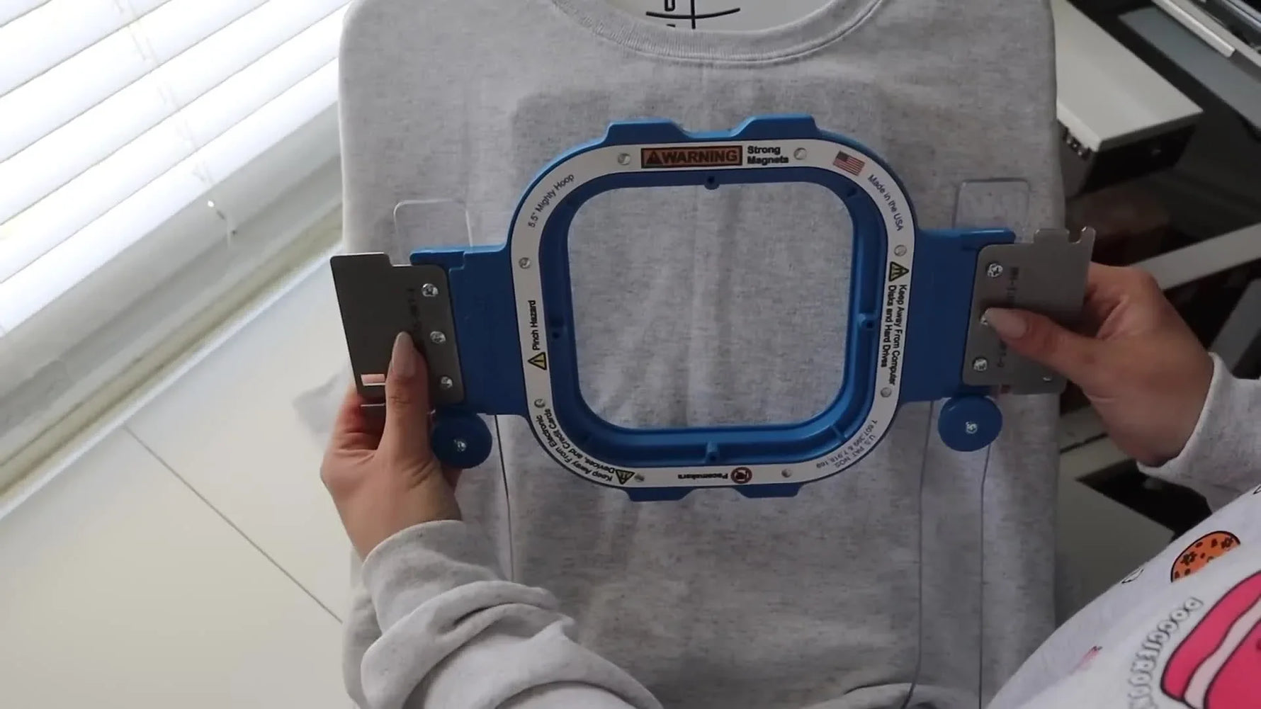 Hands snapping a blue magnetic hoop onto a sweatshirt on a hooping station, capturing the moment the magnets engage for fast, consistent hooping.