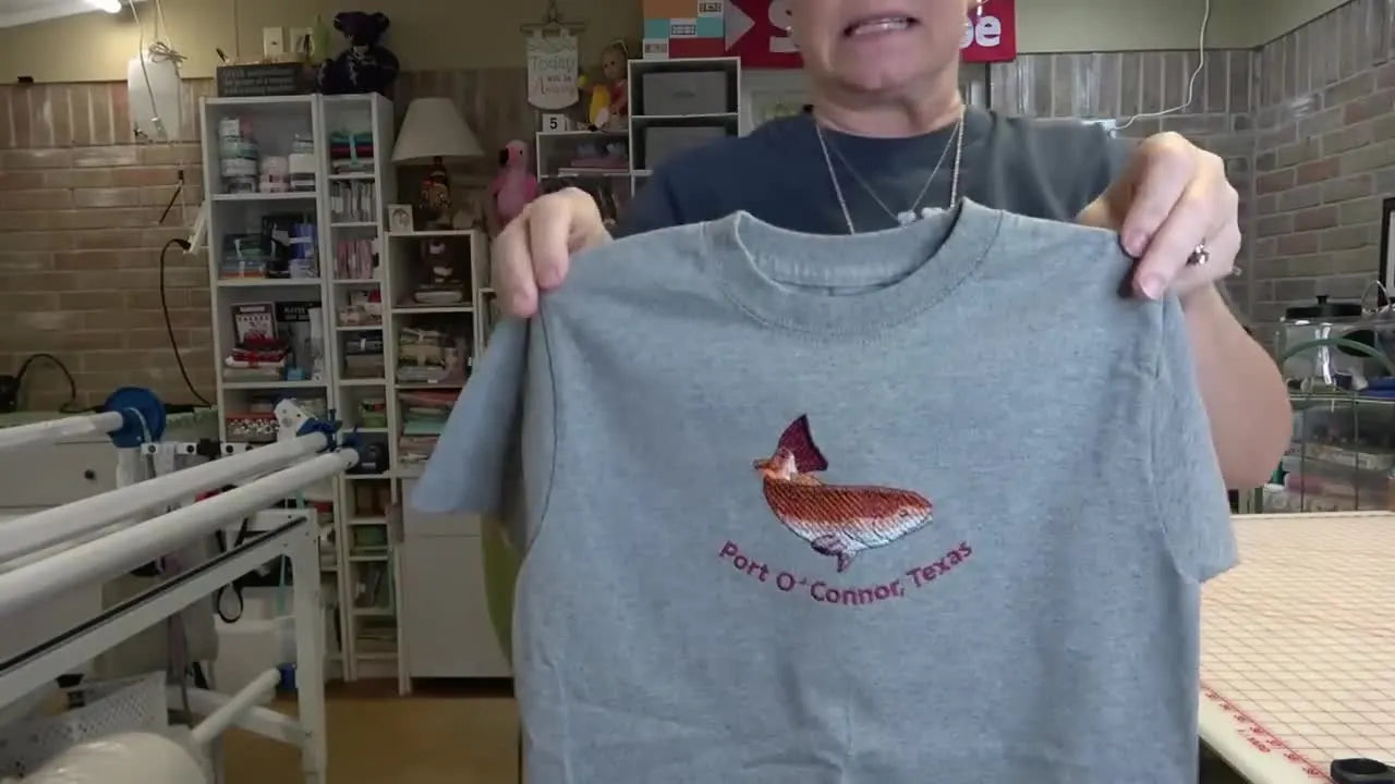 A finished grey t-shirt is held up, showing a red fish embroidery with neatly curved text stitched underneath.