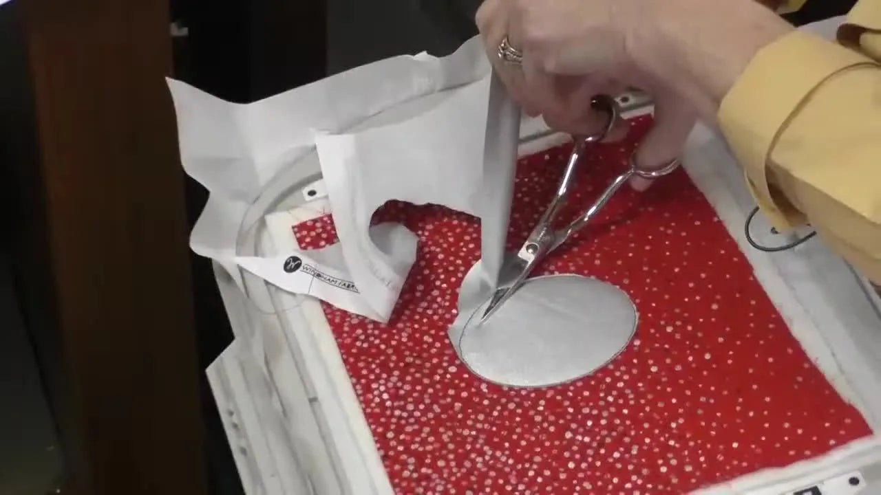 Close-up of trimming metallic appliqué fabric with duckbill scissors while the panel remains hooped for maximum stability.