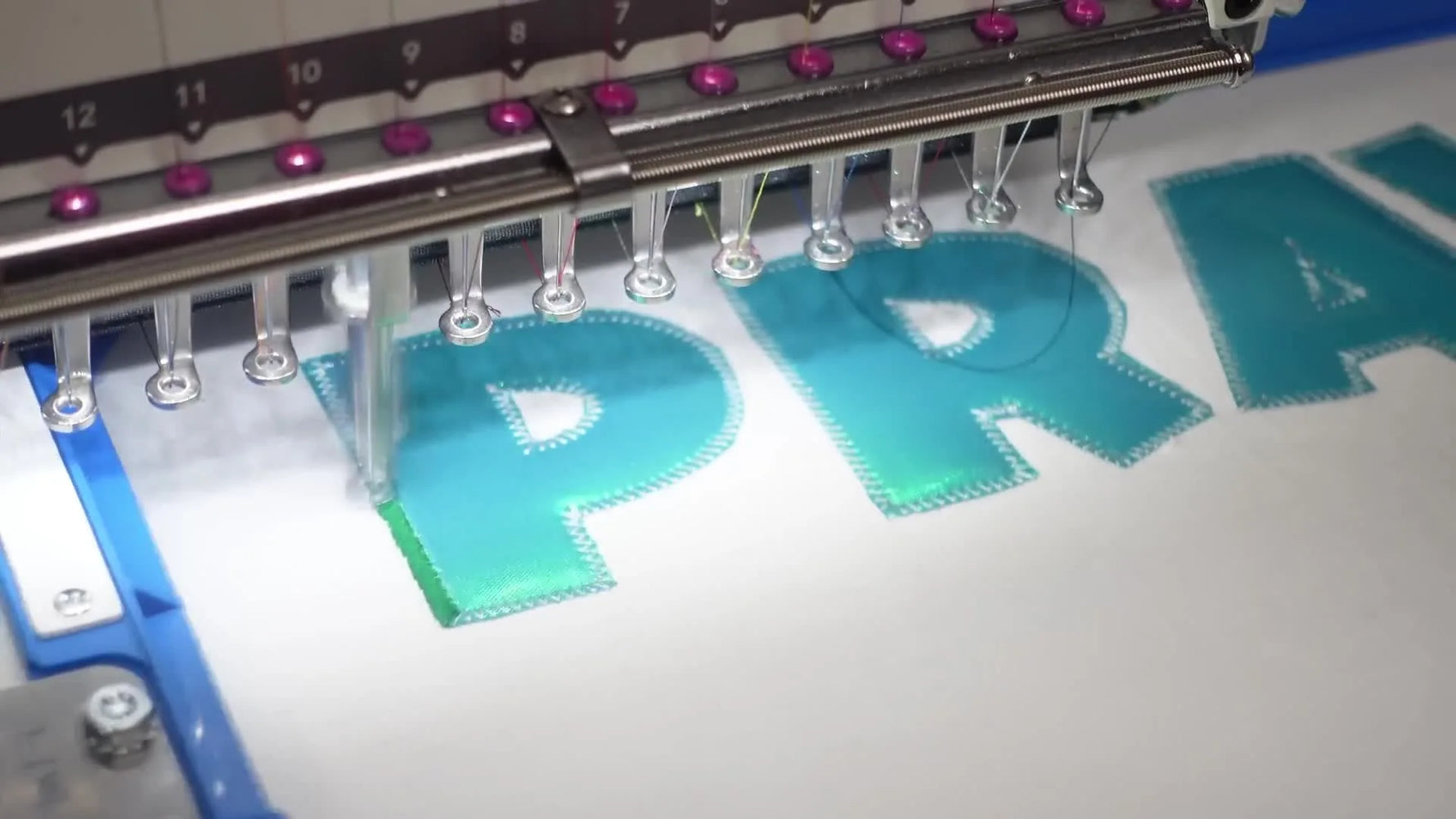 A close-up of the Bai Mirror stitching a dense satin border around teal Siser Aurora HTV appliqué letters on a hooped white T-shirt.