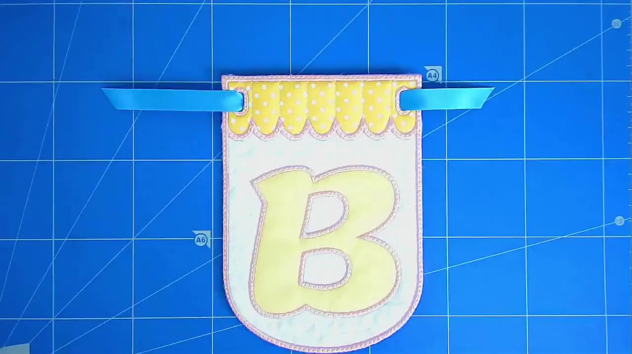 A finished in-the-hoop bunting flag with ribbon threaded through stitched eyelets, shown as a clean flat-lay reveal.
