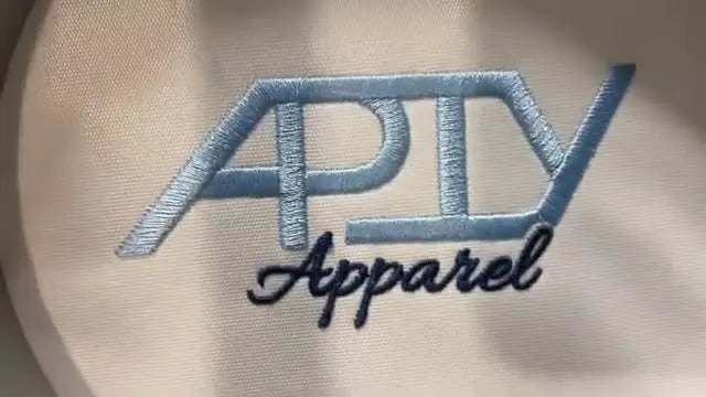 A finished two-color “APTV Apparel” logo stitched cleanly on white fabric after proper hoop selection, laser tracing, and speed control on a HappyJapan HCH Plus.