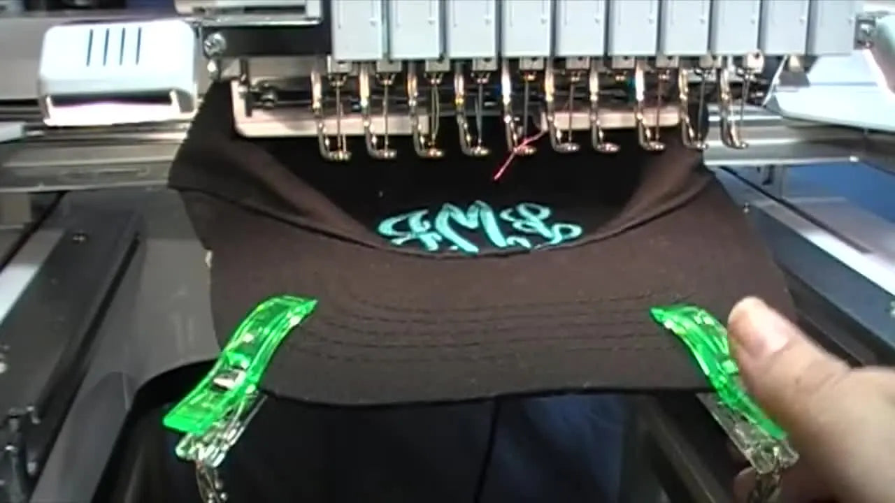 A close view of green clips and a strap holding a hanging weight that pulls a cap brim down to prevent flapping during embroidery.