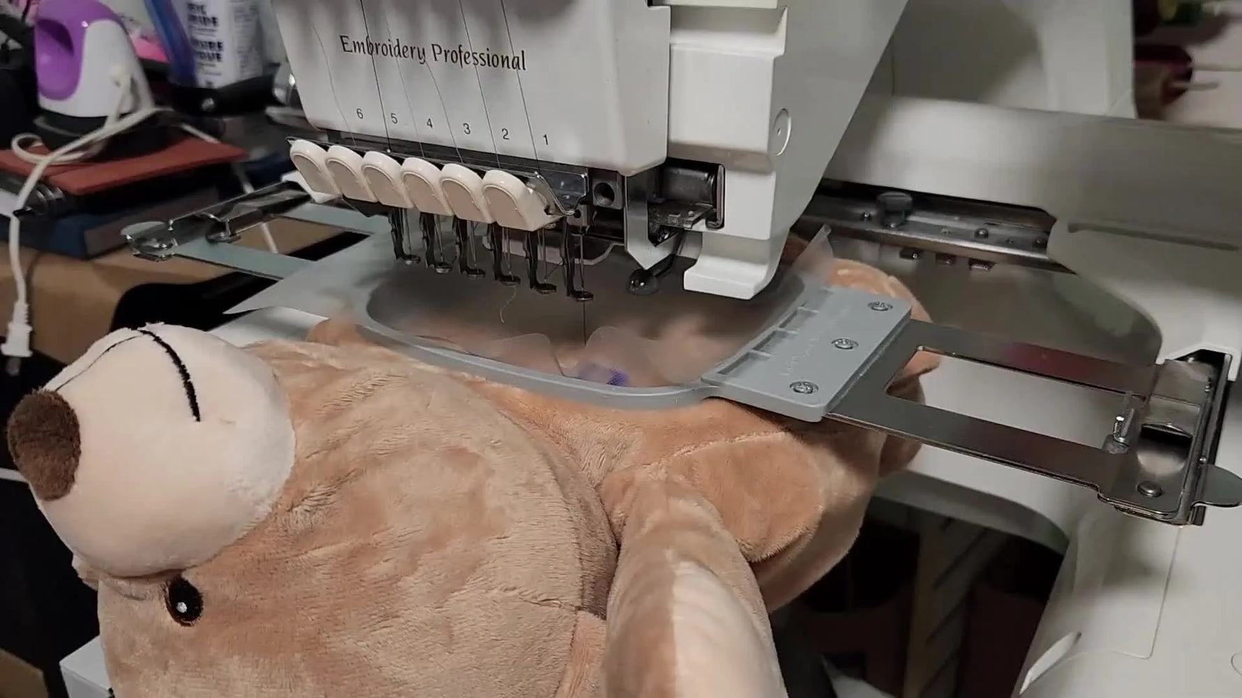 A close-up shows crisp embroidered text stitching cleanly on plush teddy bear fabric mid-process, with the topping controlling fuzz for sharp lettering.