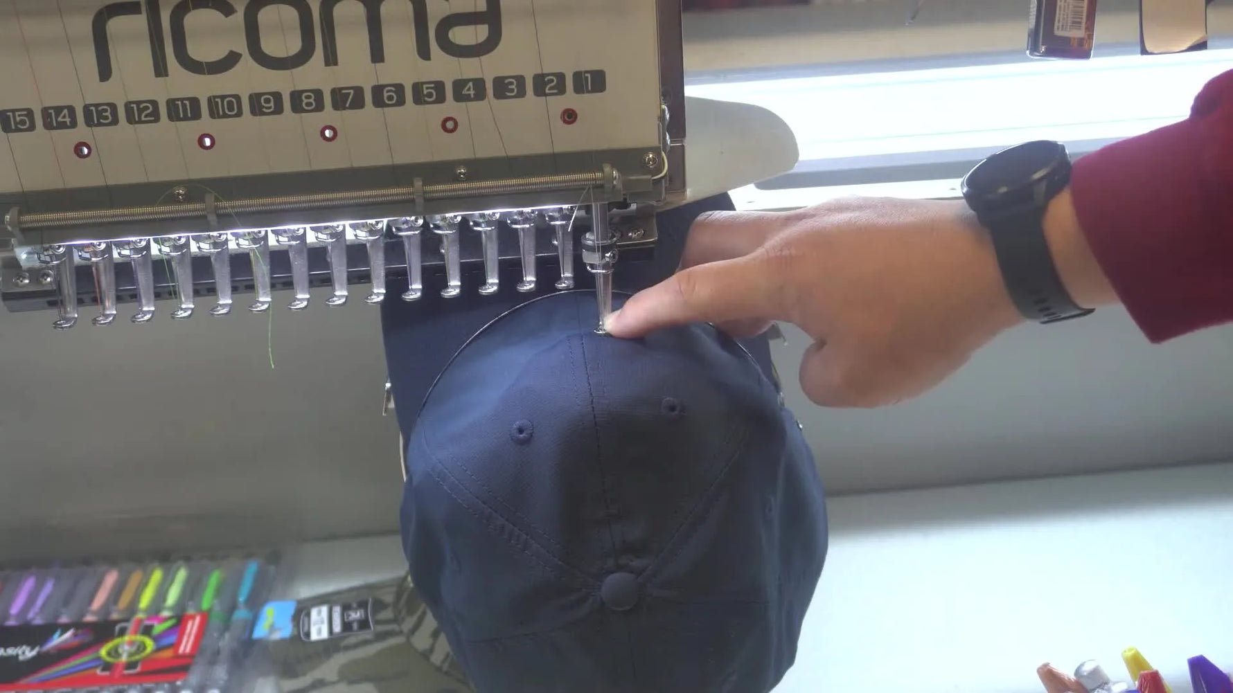 Hands rotate an unstructured dad hat on the mounted cap driver to re-center the seam under needle #1 without re-hooping.