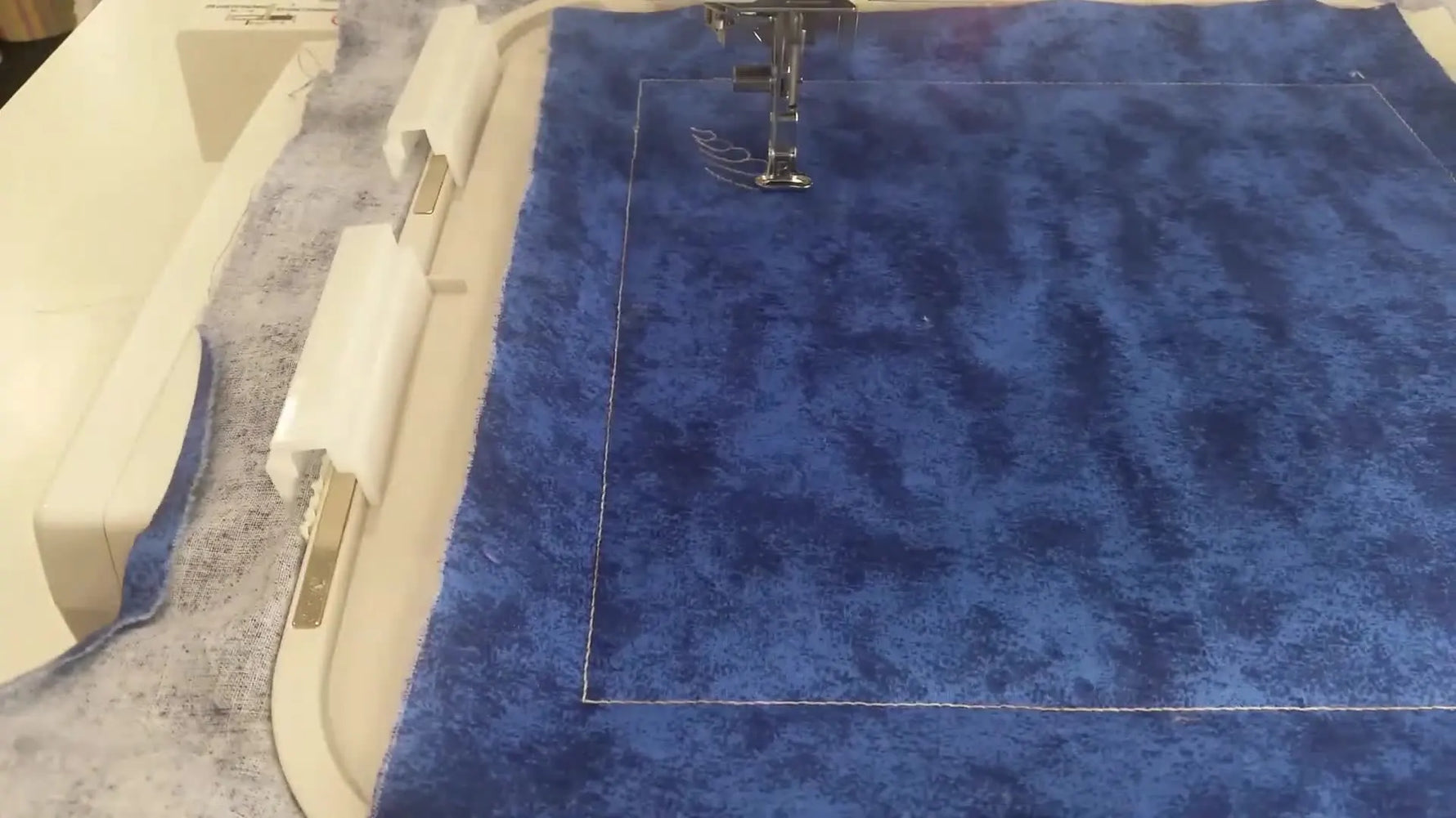 Close-up of an embroidery machine stitching a delicate diagonal feather quilting motif in light thread on blue fabric inside a large magnetic hoop.