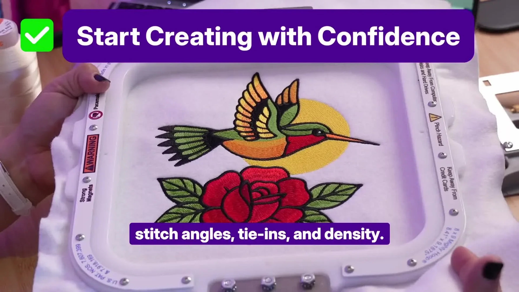 Hands hold a square magnetic hoop framing a crisp hummingbird embroidery, with the magnets visible along the frame edge.