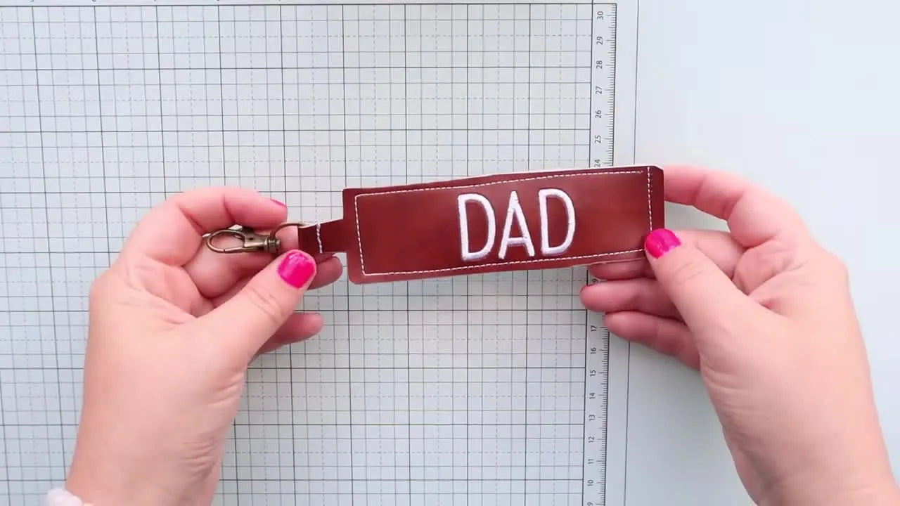 A finished faux leather keychain with crisp white “DAD” embroidery is held close to the camera, showing clean edges and a professional stitch-out.