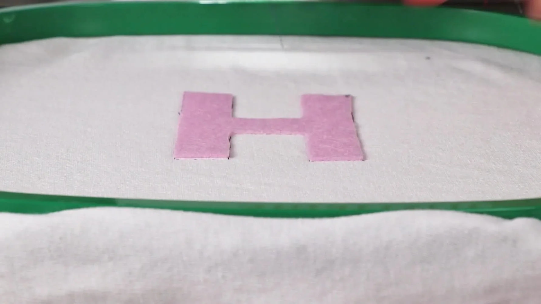 A top-down view shows the pink pre-cut “H” appliqué perfectly aligned inside the stitched placement outline on the hooped white base fabric.