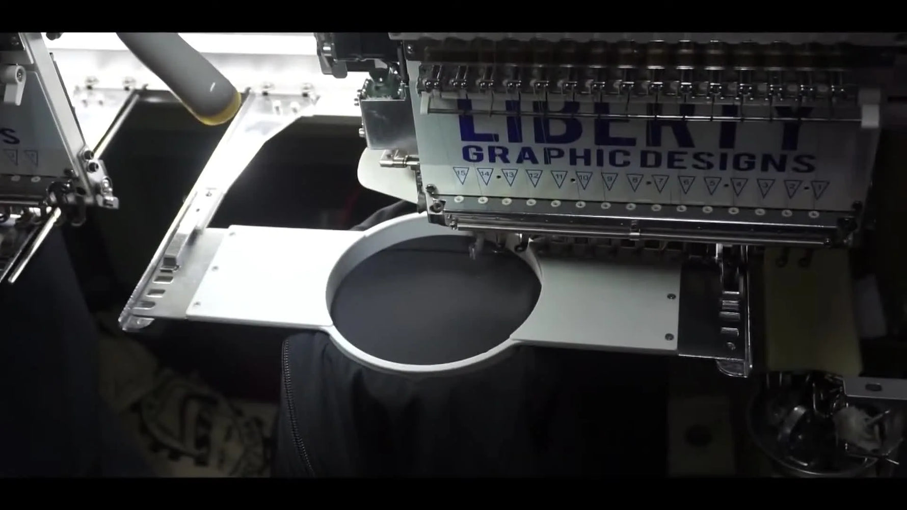 A close-up of the embroidery head actively stitching the Savage Council logo onto a black softshell jacket, showing the needle bar, presser foot, and clean stitch formation.