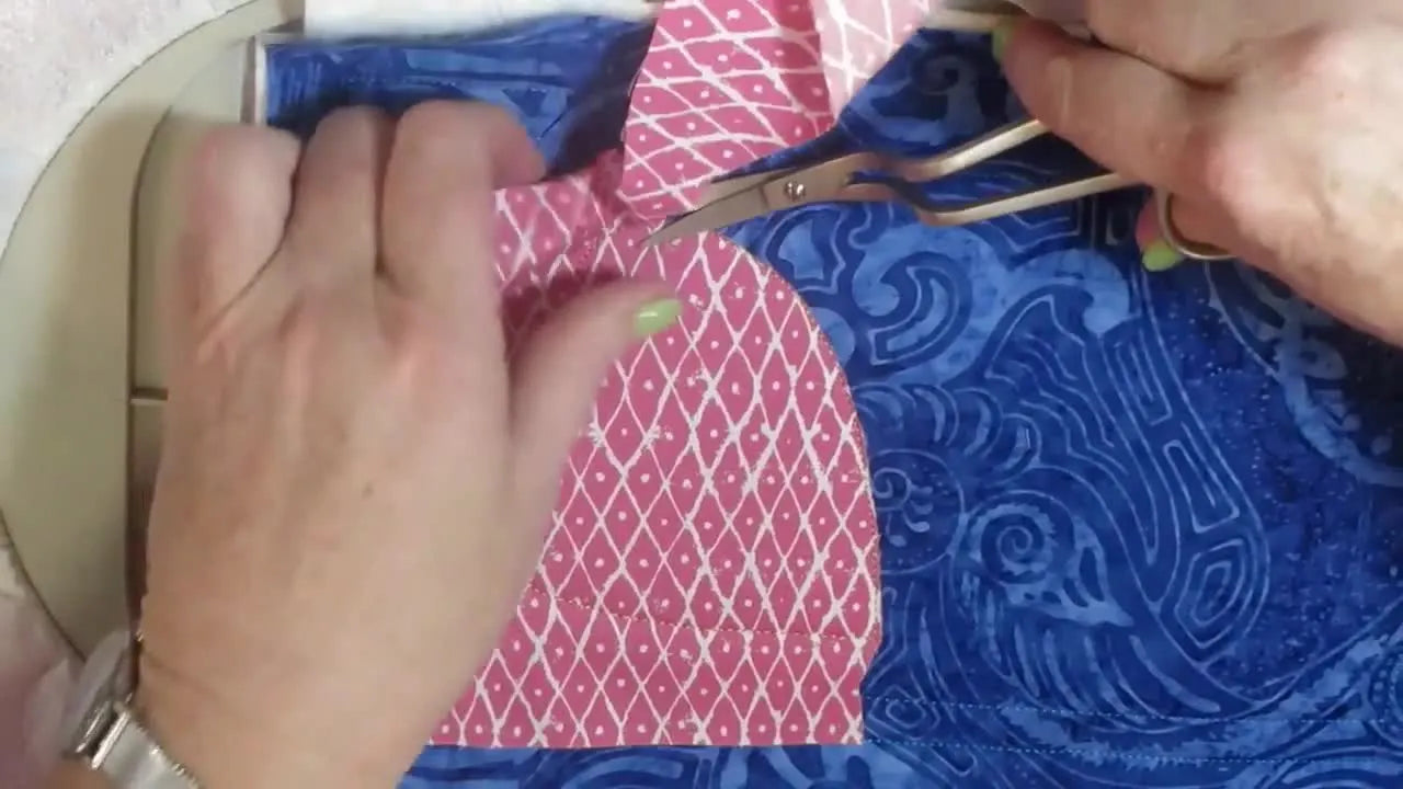 A hand uses curved embroidery scissors to trim the appliqué fabric, holding the excess fabric taut.