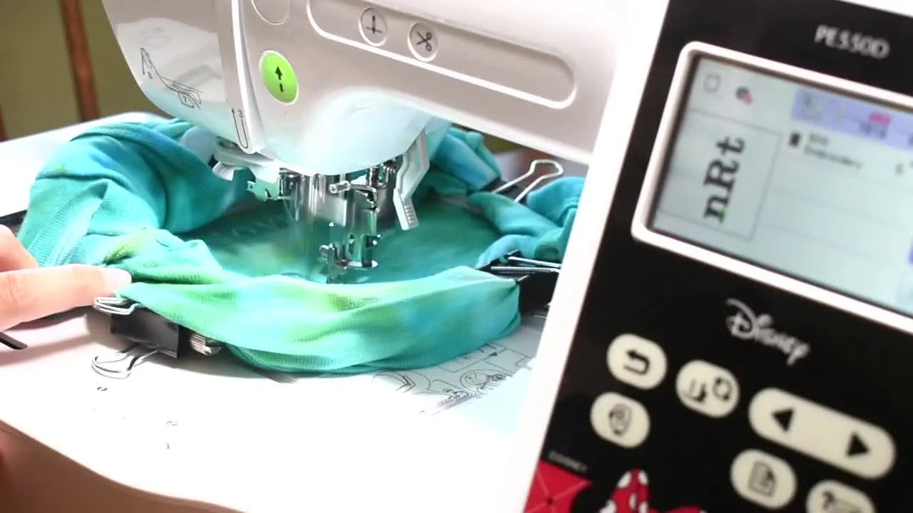 The Brother PE550D stitches teal lettering on a hooped green baby onesie while hands keep bulky fabric safely out of the moving hoop path.