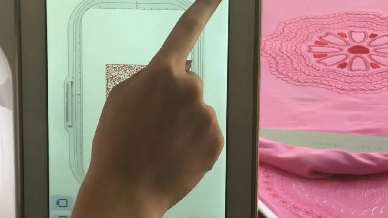 A split-screen view shows the IQ Designer artwork on the Solaris display while the machine stitches the same design onto pink fabric.