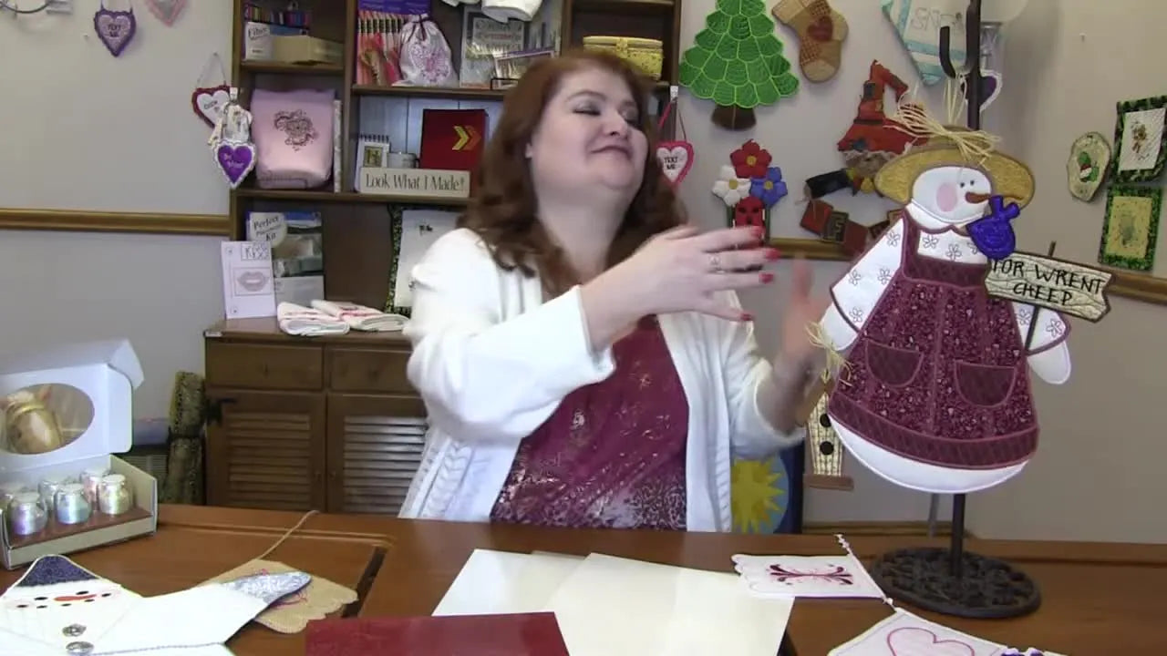 A presenter holds up the finished Garden Snow Lady in-the-hoop embroidery, highlighting the mixed-media sparkle and layered construction.
