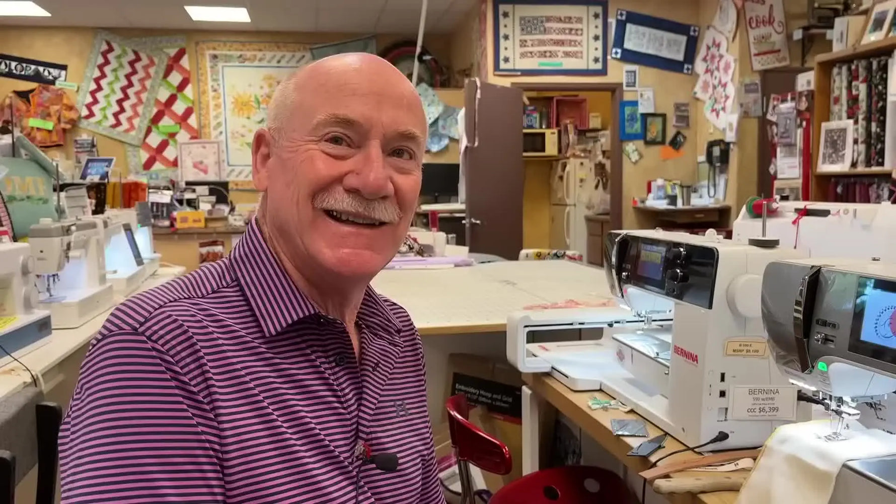 Man in a pink striped shirt talking to the camera in a sewing machine shop.