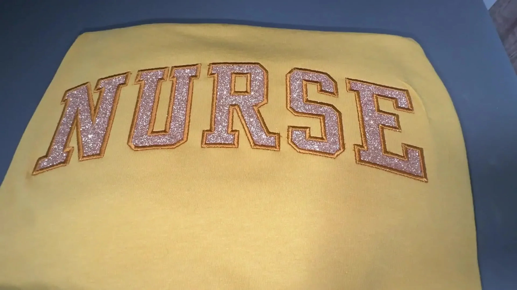 A close-up of the finished sparkling “NURSE” glitter HTV appliqué on a mustard shirt after stitching and heat pressing.