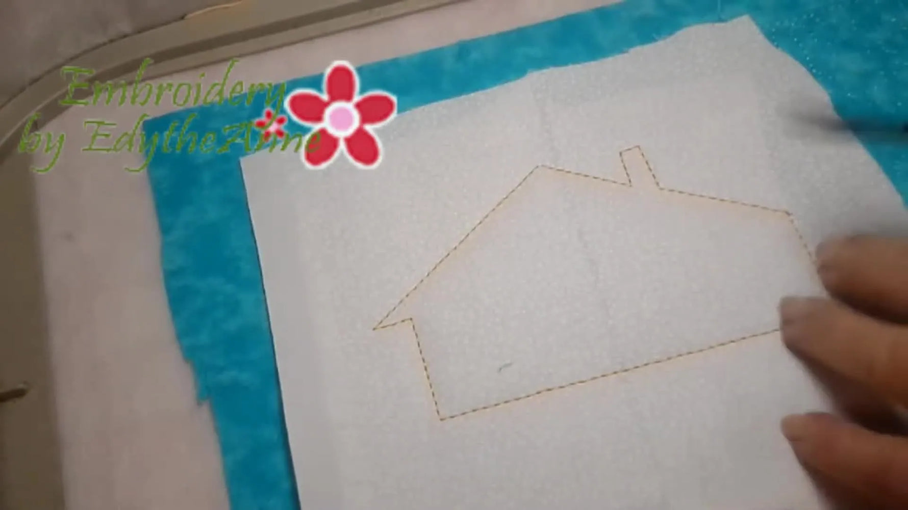 A close-up shows double-curved scissors positioned inside the hoop to begin trimming the white house appliqué cleanly along the tack-down line.