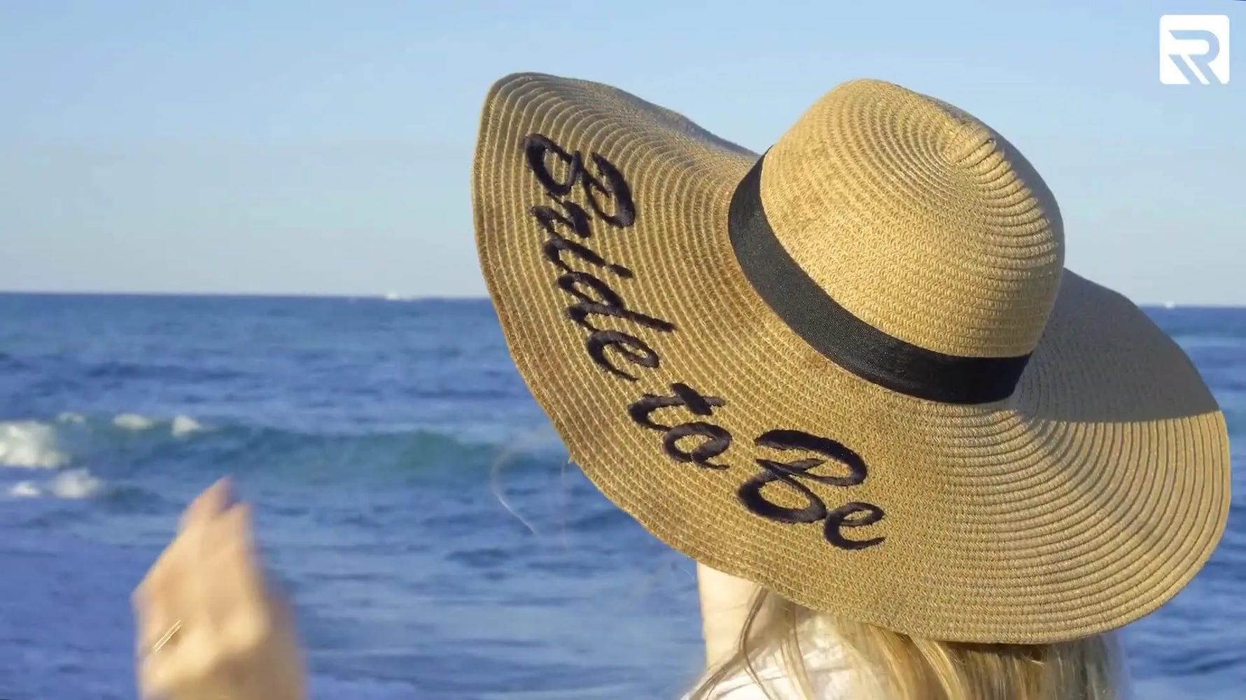 A finished wide-brim straw bridal hat sits on sand with crisp black “Bride to Be” embroidery visible on the brim.