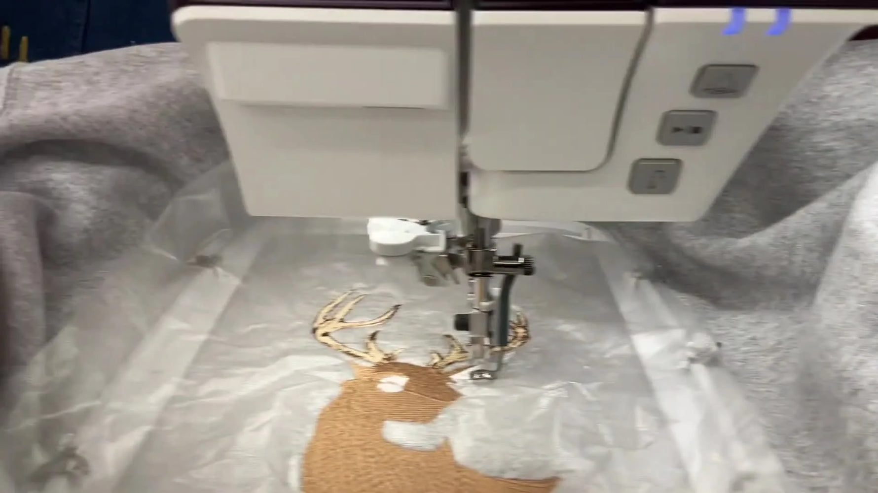 A close-up of the Pfaff Creative Icon 2 stitching a realistic deer on a grey fleece sweatshirt with water-soluble topping visible on the surface.
