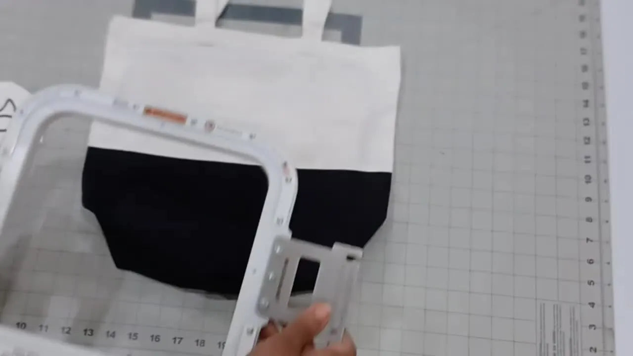 Hands lower the top magnetic frame onto a hooped tote bag at the hooping station, moments before the magnets snap into place.