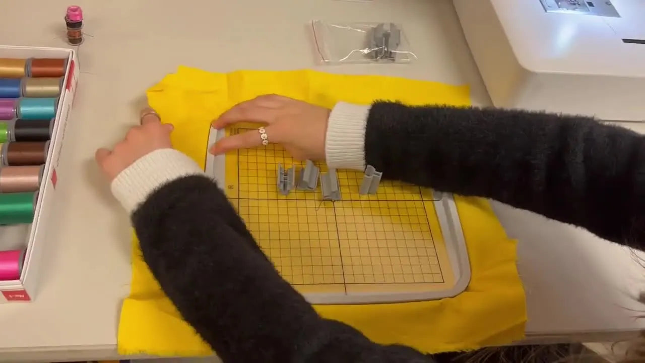 Close-up of grey hoop clips clamping fabric tightly around a Janome 200x200 hoop for stable, wrinkle-free stitching.