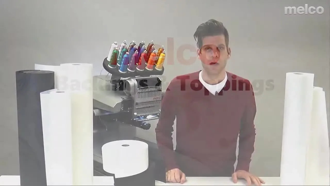 A presenter stands beside a Melco multi-needle embroidery machine with multiple stabilizer rolls laid out for comparison.