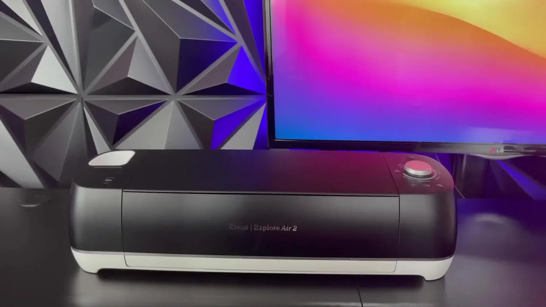A black Cricut Explore Air 2 machine sits on a desk with a geometric wall and colorful screen in the background.