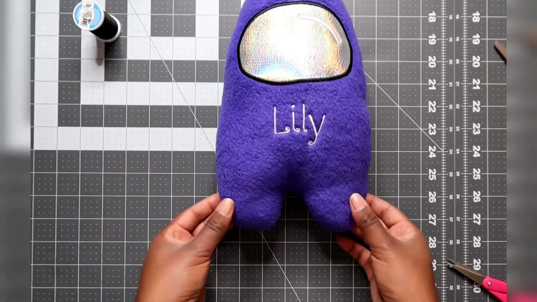 A finished purple crewmate plushie stands upright with holographic vinyl goggles, showing crisp applique edges and a clean, nearly invisible bottom closure.