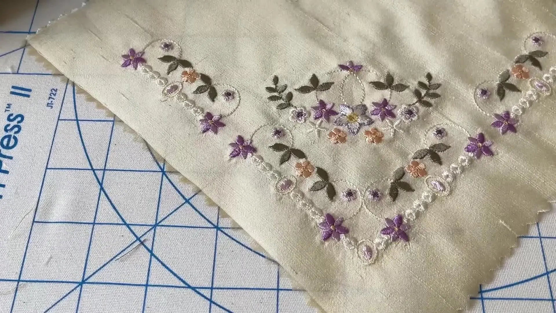 The finished corner embroidery on silk lies flat on a gridded cutting mat, showing the completed placement and clean edges after pressing.
