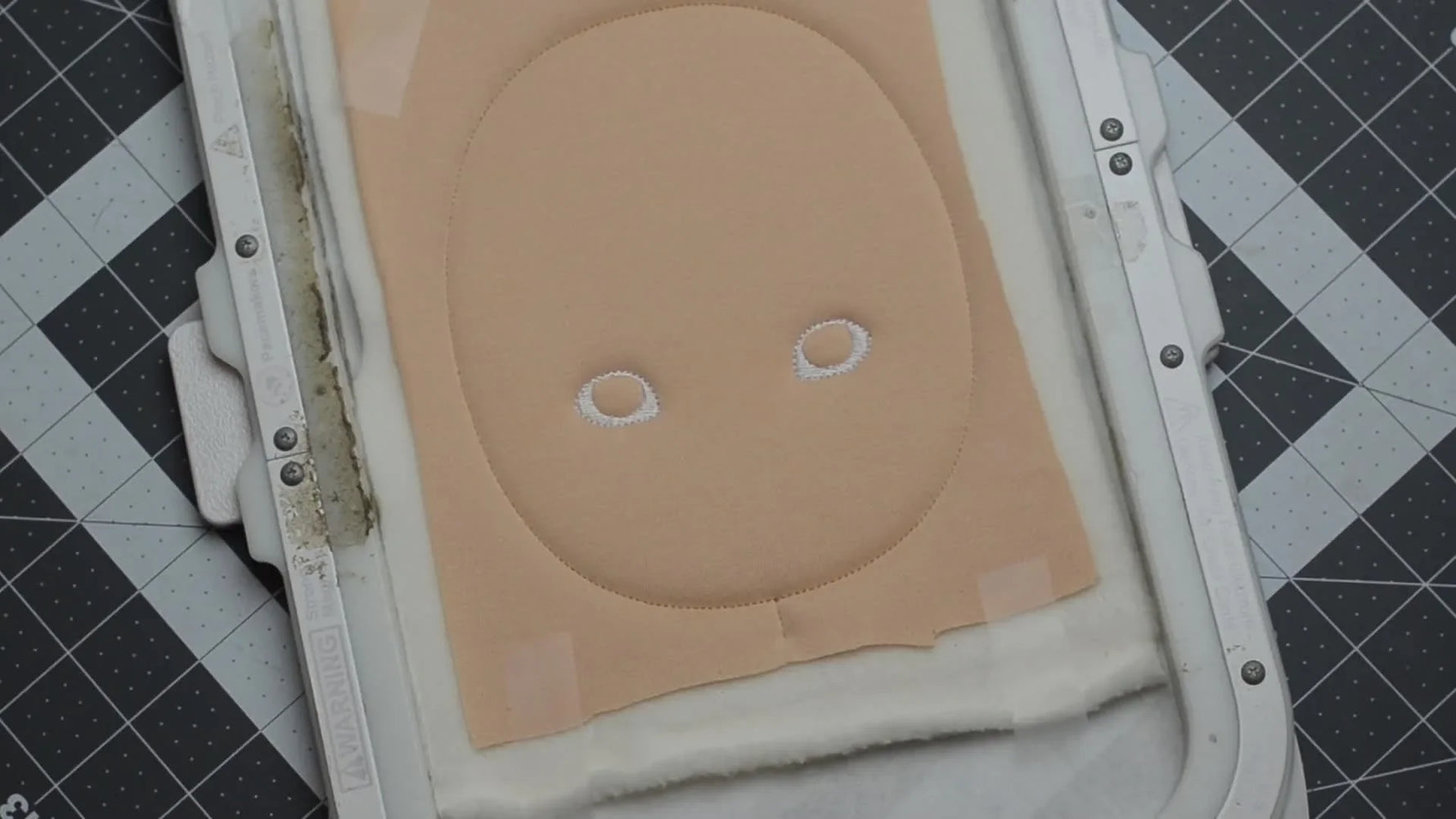 A close-up of the machine finishing the doll’s embroidered eyes in the hoop, showing dense facial stitching on jersey stabilized for a clean result.
