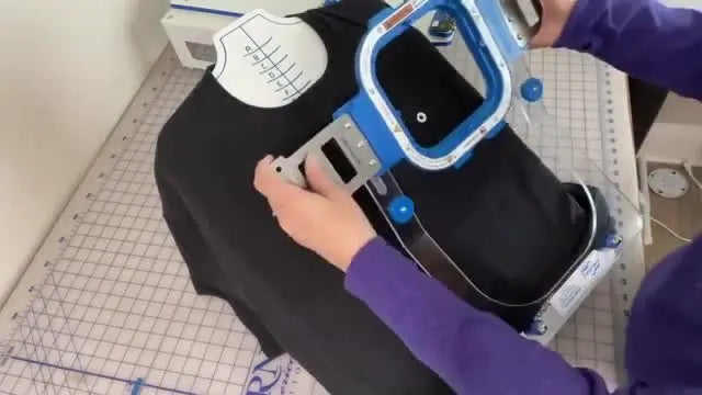 A close-up shows the top magnetic hoop with the warning label visible, moments before it snaps into place over a left-chest t-shirt setup on the station.