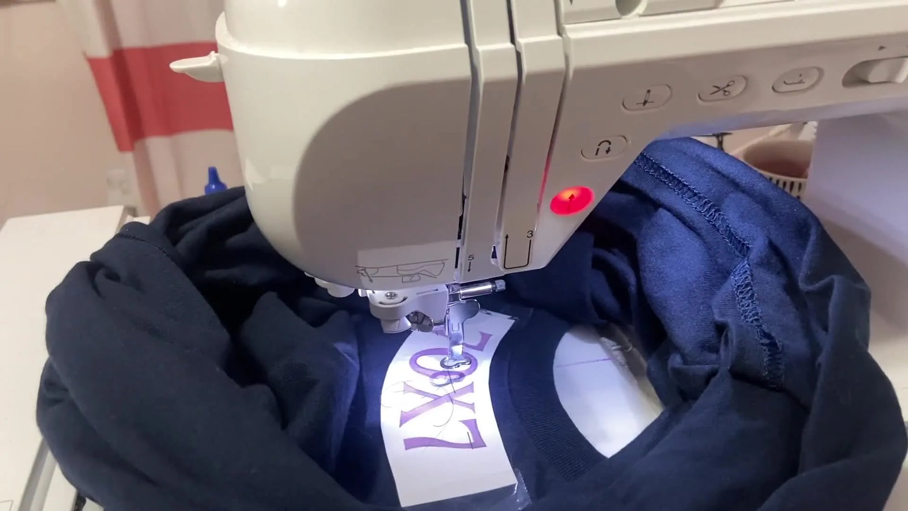 A close-up of a Brother single-needle machine stitching white thread into a blue sweatshirt neckline, showing the curved text placement in progress.