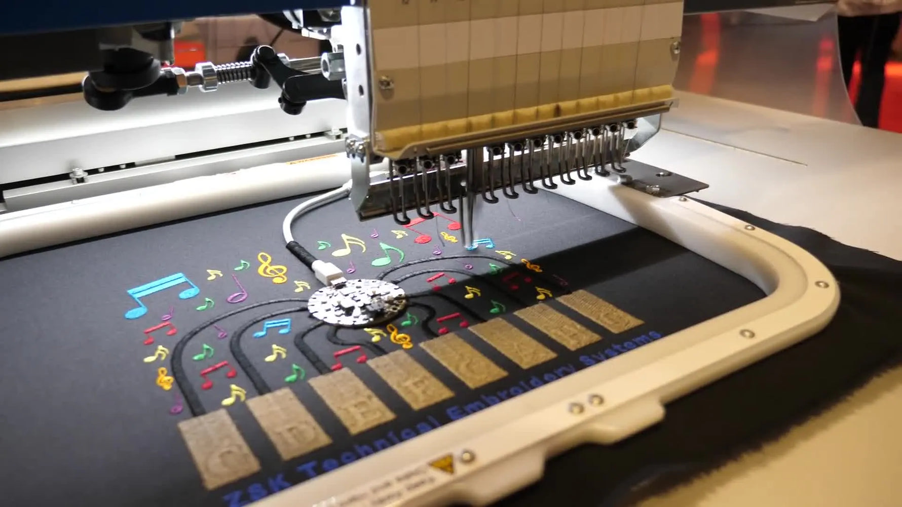 A close-up of a ZSK needle bar stitching a colorful technical embroidery design on black fabric during a smart-textile demo.
