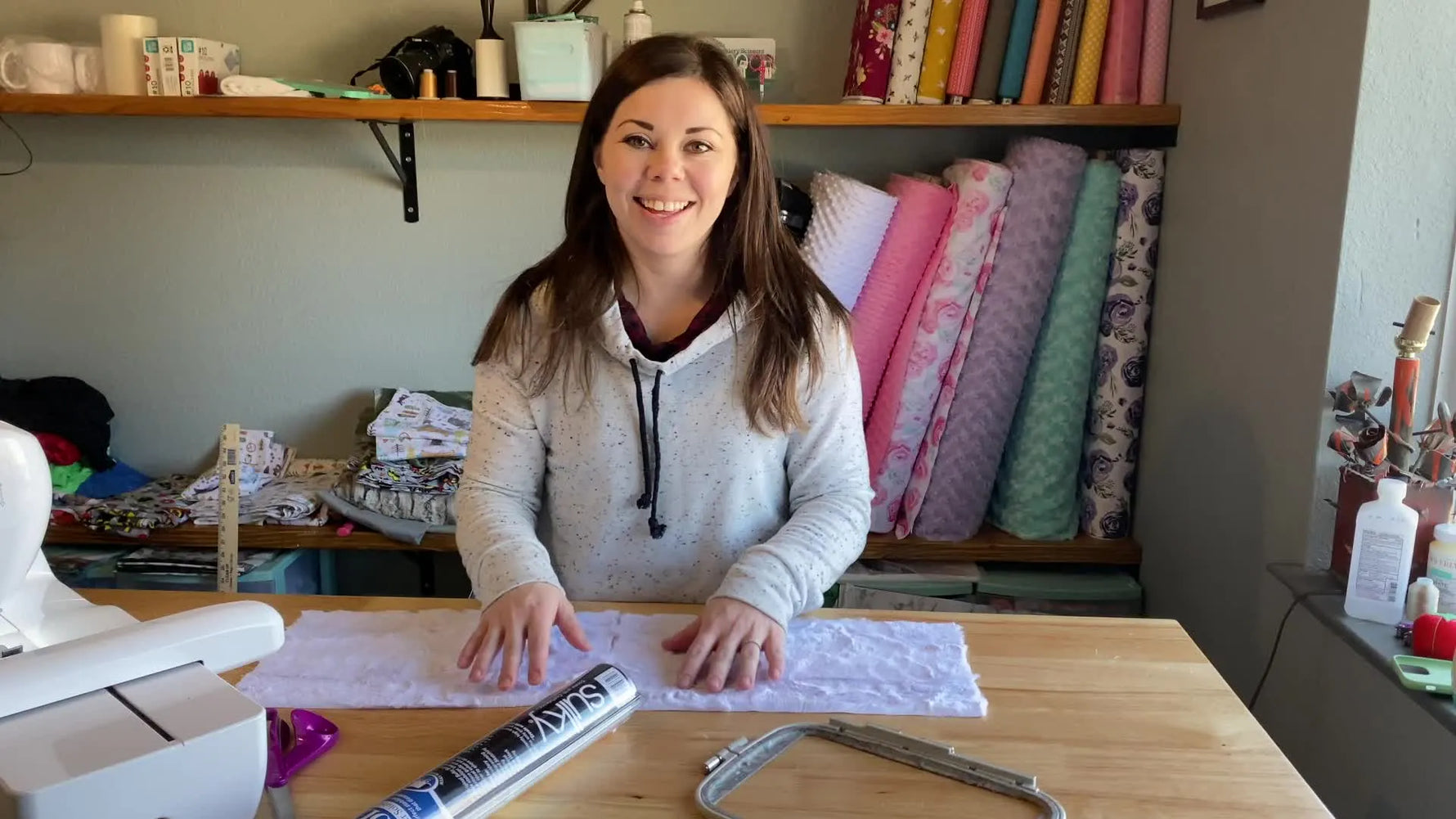 Kayla introducing the topic of water-soluble stabilizer for baby blankets, showing fabric samples on a wooden table.