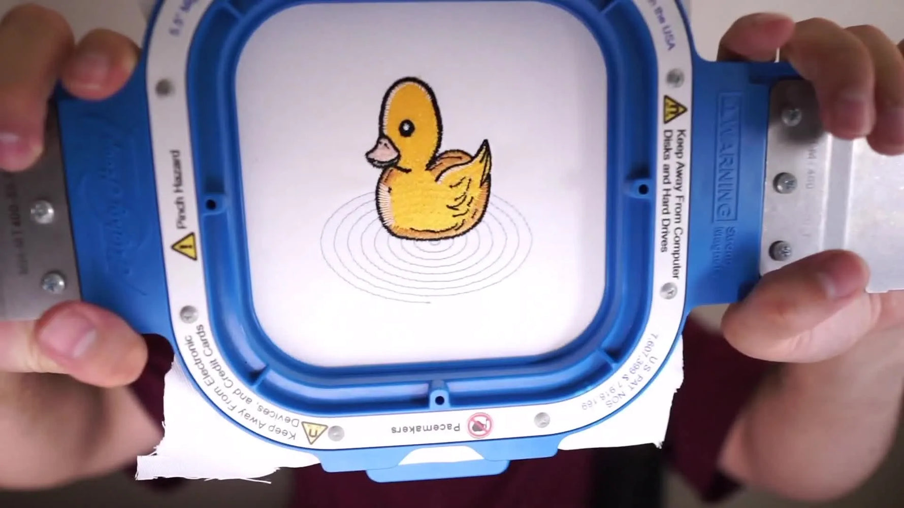 A finished yellow duck embroidery is shown stitched on white fabric and held in a blue-and-white magnetic hoop.