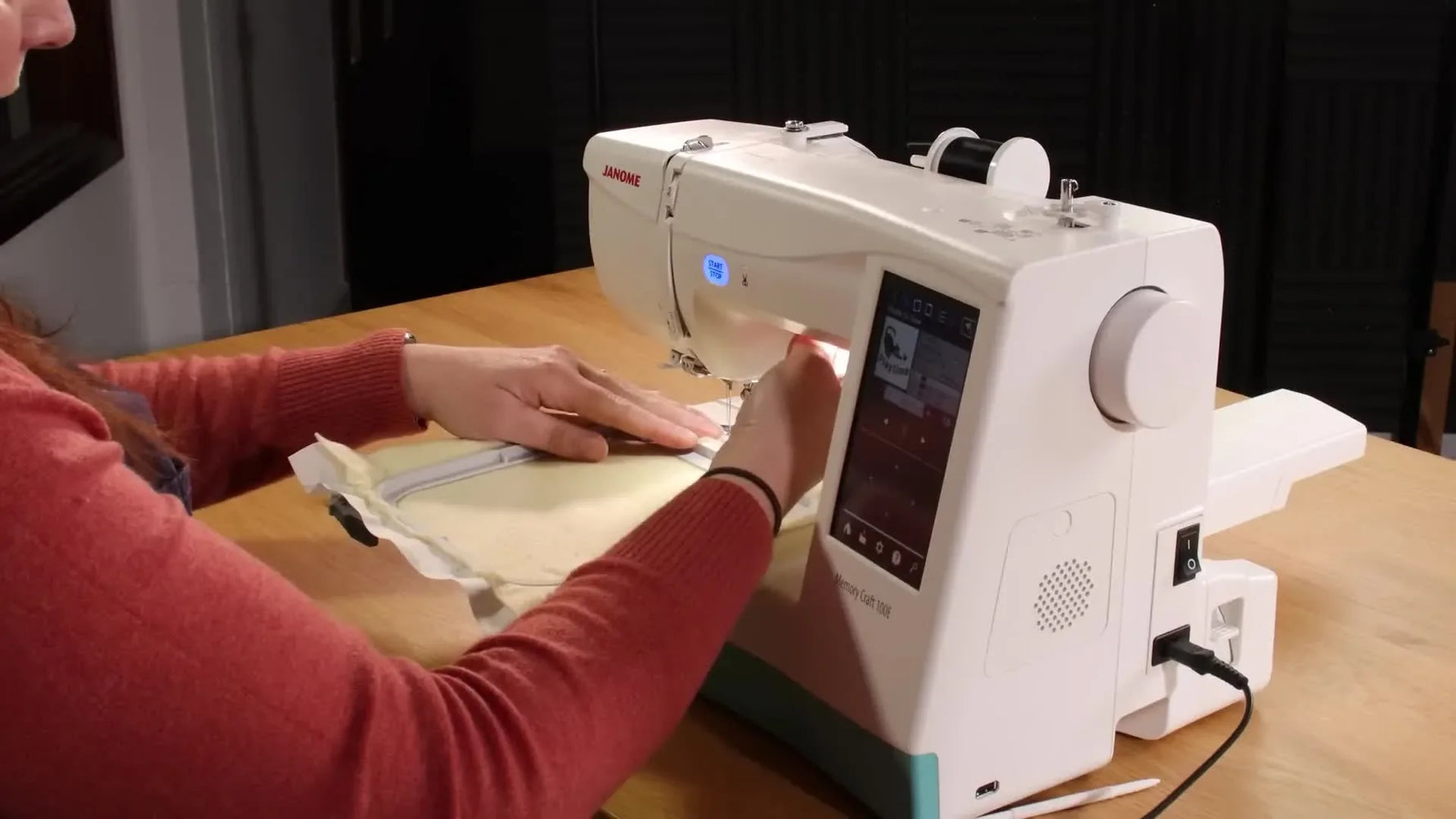 A close-up shows the hoop being attached to the Janome Memory Craft 100E embroidery arm using the lock lever, right before stitching begins.
